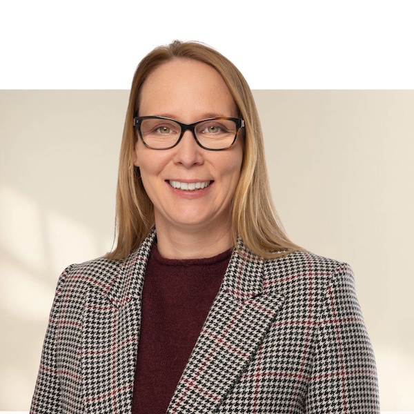 Porträt von Barbara Dietrich mit glatten, blonden Haaren, schwarzer Brille, karierter Jacke in Schwarz-Weiss-Rot und bordeauxfarbenem Oberteil vor unscharfem Hintergrund