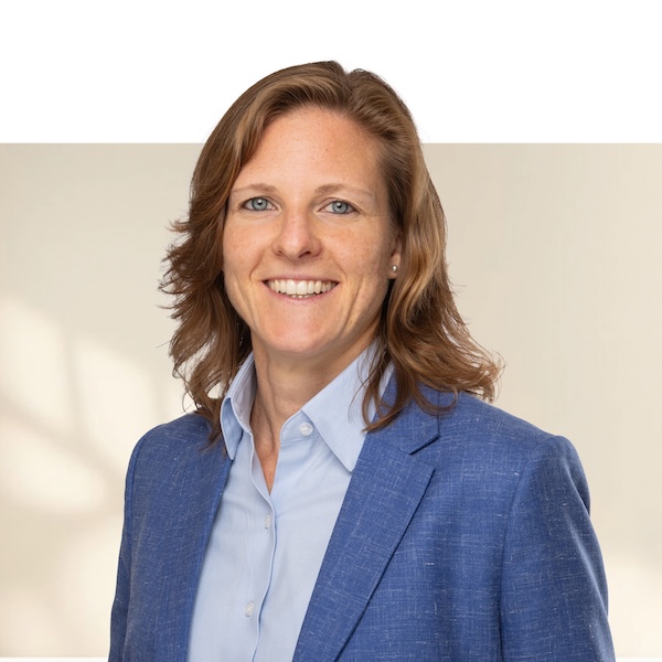 Porträt von Claudia Bachmann mit mittellangen, gewellten, rötlich-blonden Haaren, blauem Blazer und hellem Hemd vor neutralem, unscharfem Hintergrund