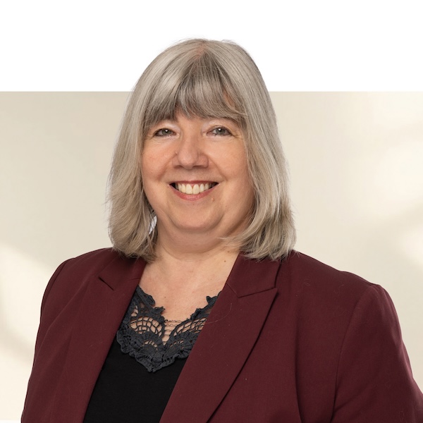 Porträt von Evelyne Steiner mit grauen, kinnlangen Haaren, bordeauxrotem Blazer und schwarzem Spitzentop vor unscharfem Hintergrund