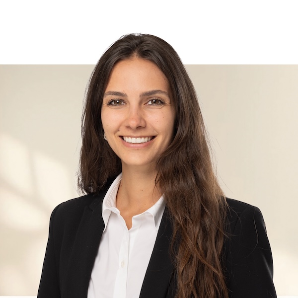 Porträt von Jenny Styger mit langen, braunen Haaren, schwarzem Blazer und weissem Hemd vor unscharfem Hintergrund