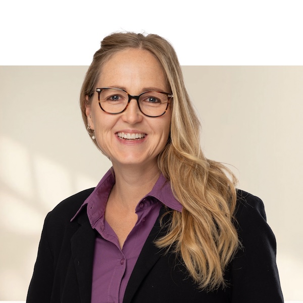 Porträt von Katrin Scheu mit langen, blonden Haaren, violetter Bluse, schwarzem Blazer und Brille vor neutralem, unscharfem Hintergrund