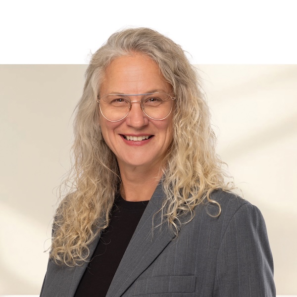 Porträt von Petra Senija mit langen, lockigen, graublonden Haaren, runder Brille und grauem Blazer vor neutralem, unscharfem Hintergrund