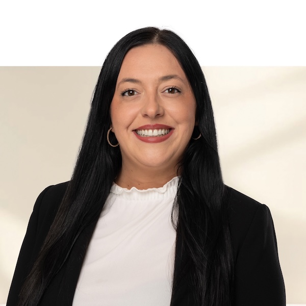 Porträt von Stephanie Frei mit langen, schwarzen Haaren, weisser Bluse mit Rüschenkragen, schwarzem Blazer und goldenen Creolen vor unscharfem Hintergrund