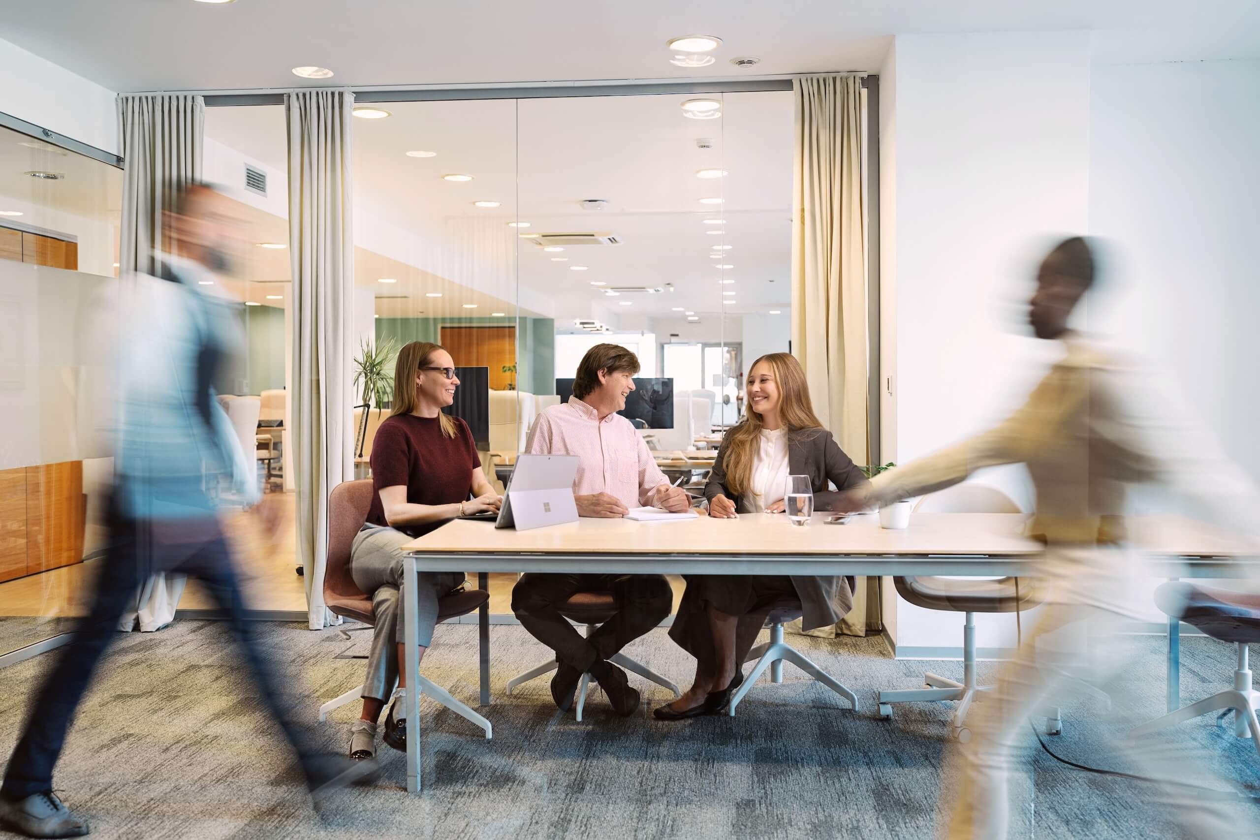 Drei Personen im Gespräch an einem modernen Konferenztisch in einem hellen Büro – im Vordergrund laufen zwei Personen in Bewegung vorbei, was dem Bild eine dynamische Atmosphäre verleiht